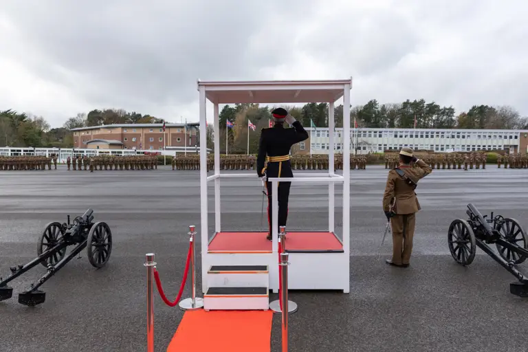 A military officer salutes from a podium with a red carpet, facing troops in formation. Two cannons are positioned on either side.