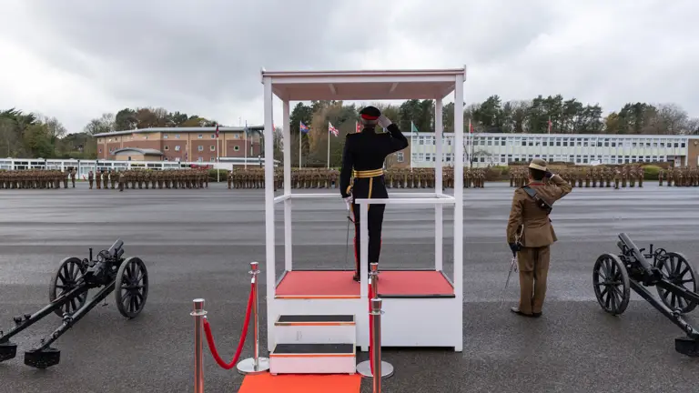 A military officer salutes from a podium with a red carpet, facing troops in formation. Two cannons are positioned on either side.
