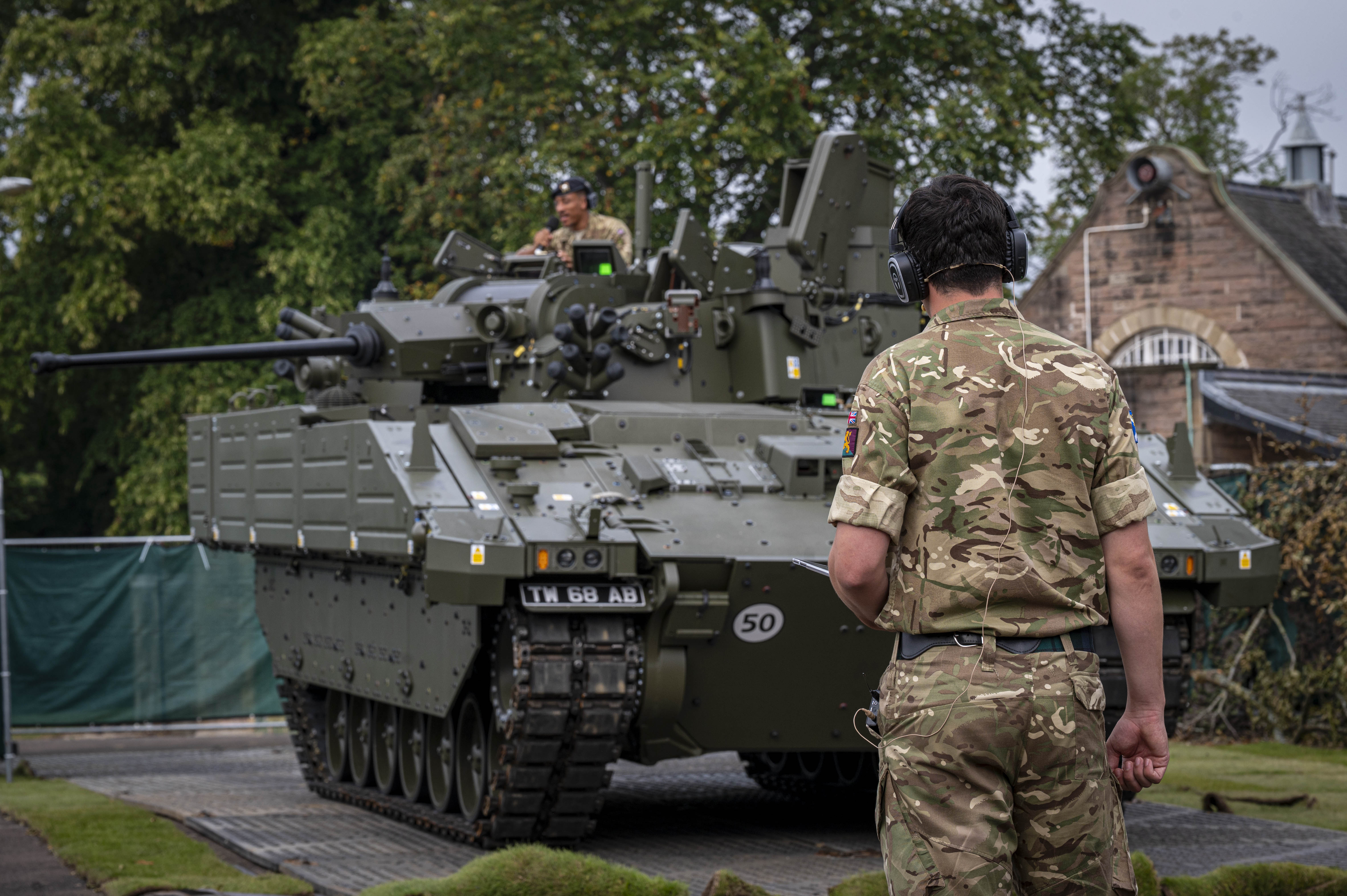 A soldier in camouflage uniform and headphones stands facing a green armored military vehicle on a paved path.
