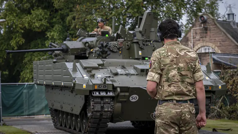 A soldier in camouflage uniform and headphones stands facing a green armored military vehicle on a paved path.