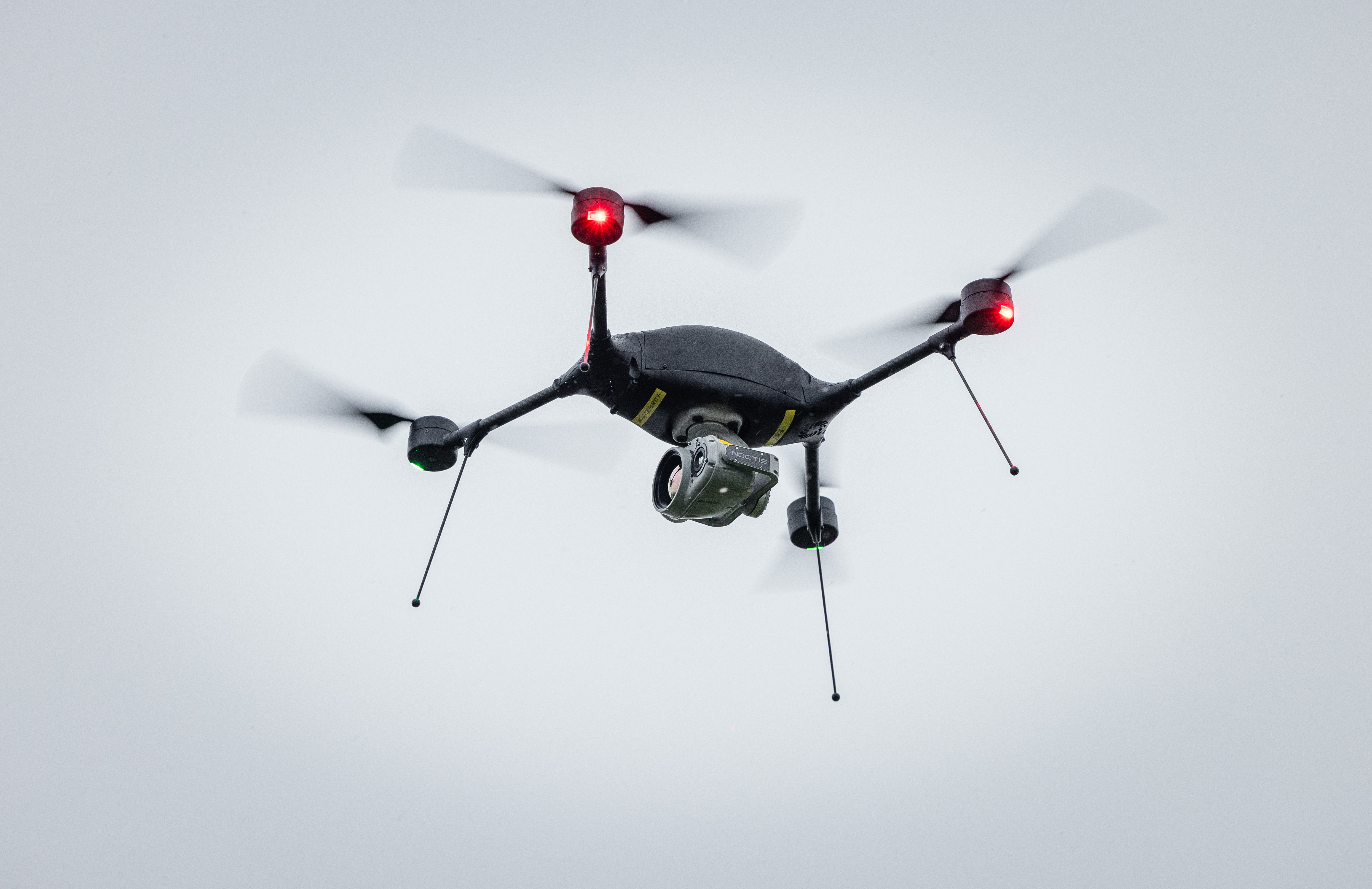 A black drone flies against a grey sky.