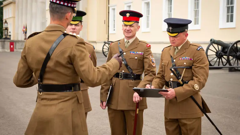 Four British Army personnel are standing in front of a large white building, with historical cannons at the base of the building.