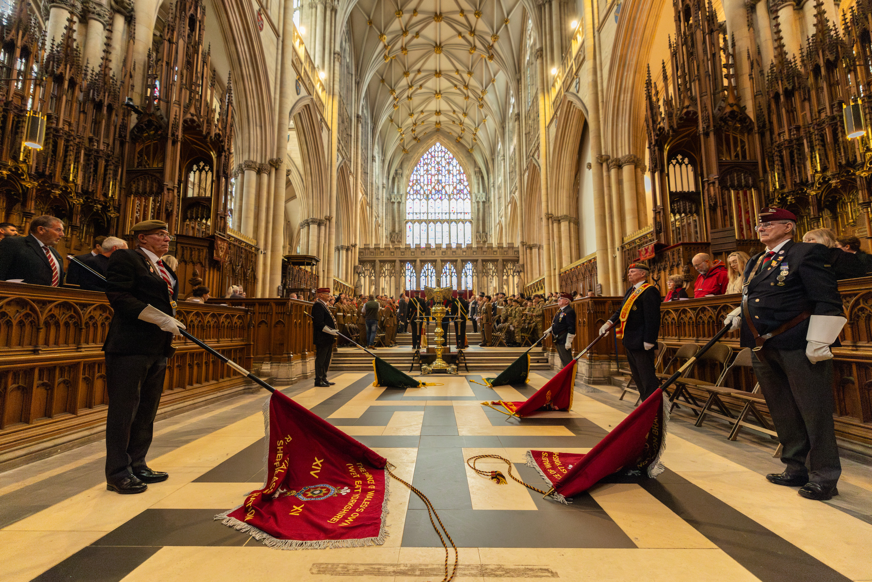 A ceremonial event is taking place inside a large, ornate cathedral. Several people are standing, holding flags that are lowered and resting on the floor. Stained glass windows are visible in the background, and guests are seated in rows along the sides. The architecture features tall arches and intricate carvings.