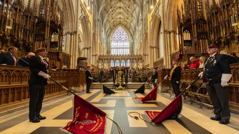A ceremonial event is taking place inside a large, ornate cathedral. Several people are standing, holding flags that are lowered and resting on the floor. Stained glass windows are visible in the background, and guests are seated in rows along the sides. The architecture features tall arches and intricate carvings.