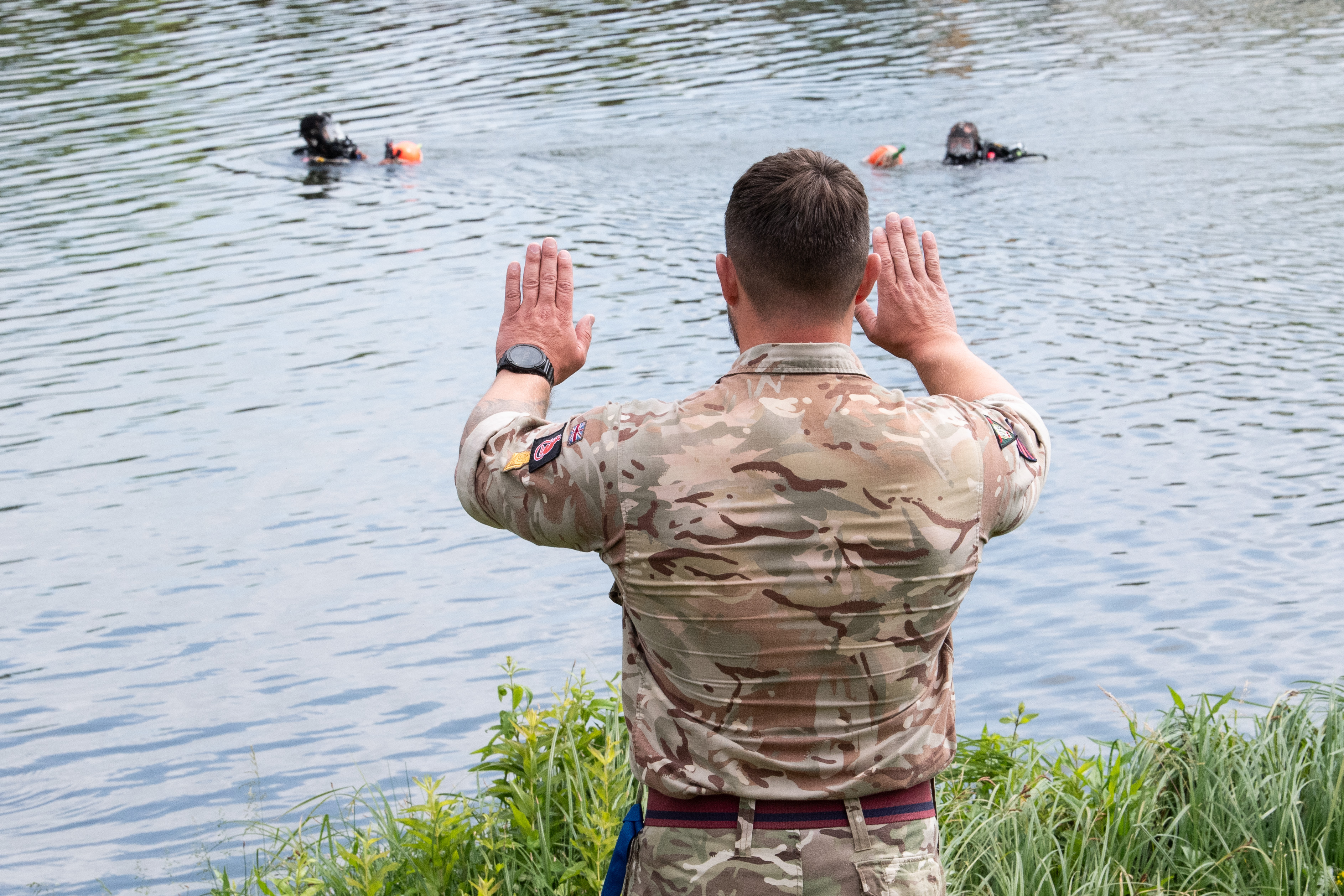 Soldier in camouflage uniform signals with raised hands to two divers in full gear floating in a lake.