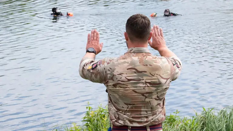 Soldier in camouflage uniform signals with raised hands to two divers in full gear floating in a lake.