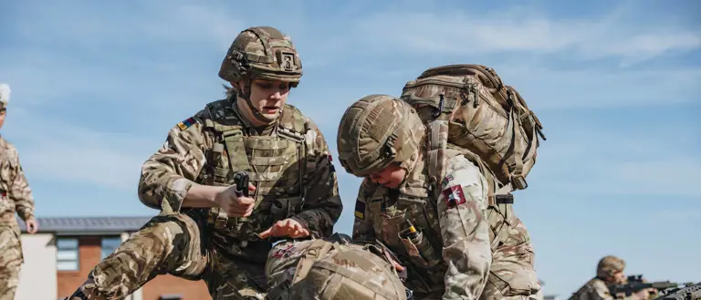 Soldiers in camouflage gear perform a medical training exercise on a grassy field under a clear blue sky.