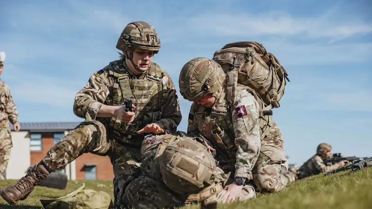 Soldiers in camouflage gear perform a medical training exercise on a grassy field under a clear blue sky.
