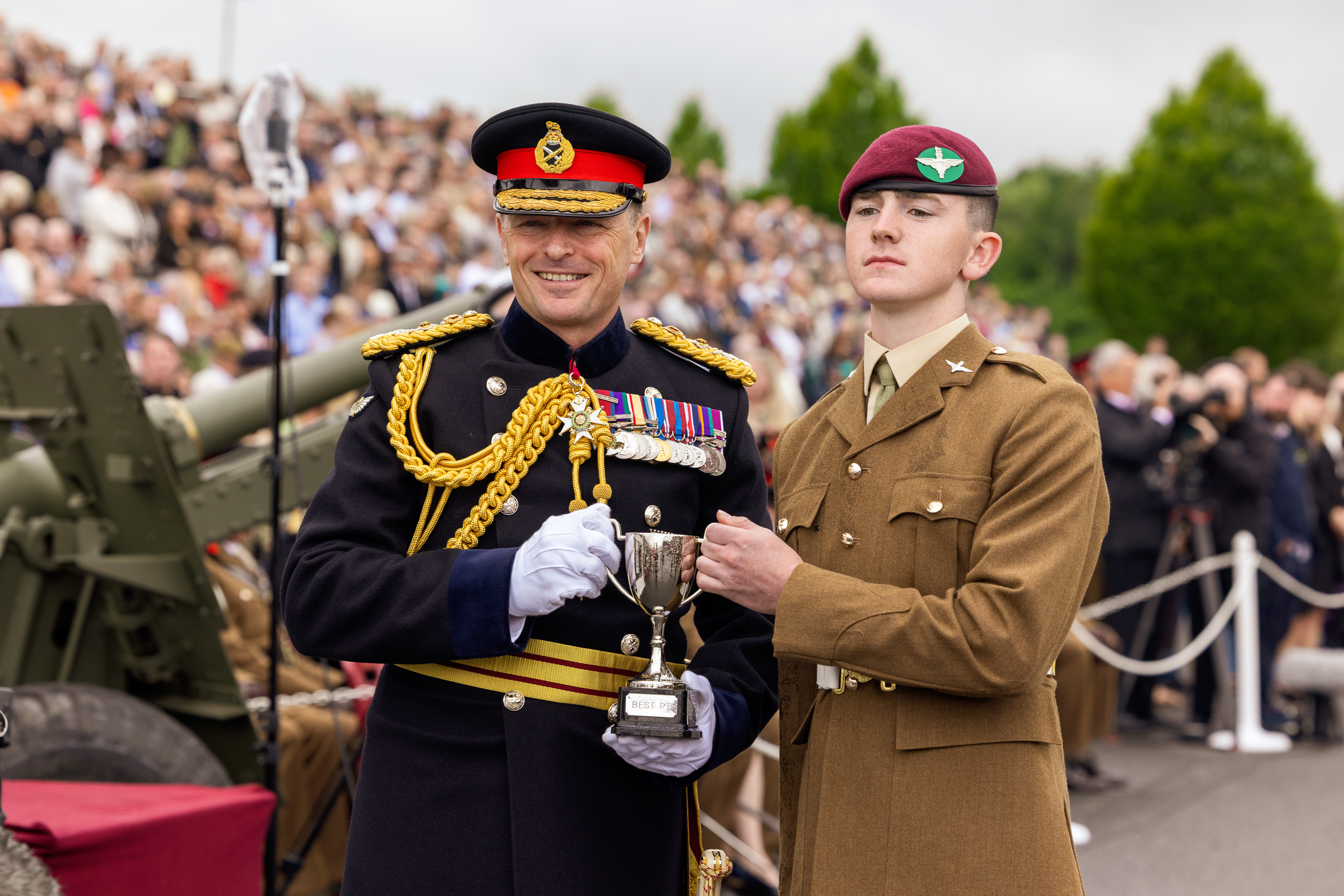 Chief of the General Staff in his blues uniform is photographed presenting a trophy award to a soldier in their brown uniform wearing a parachute regiment pink beret while smiling. 
