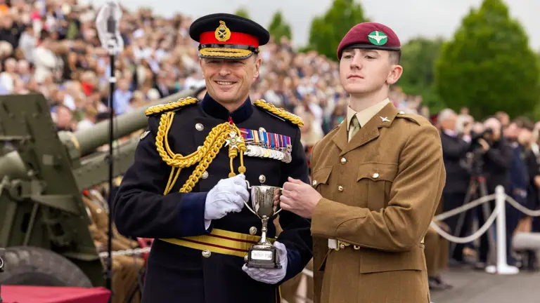 Chief of the General Staff in his blues uniform is photographed presenting a trophy award to a soldier in their brown uniform wearing a parachute regiment pink beret while smiling.