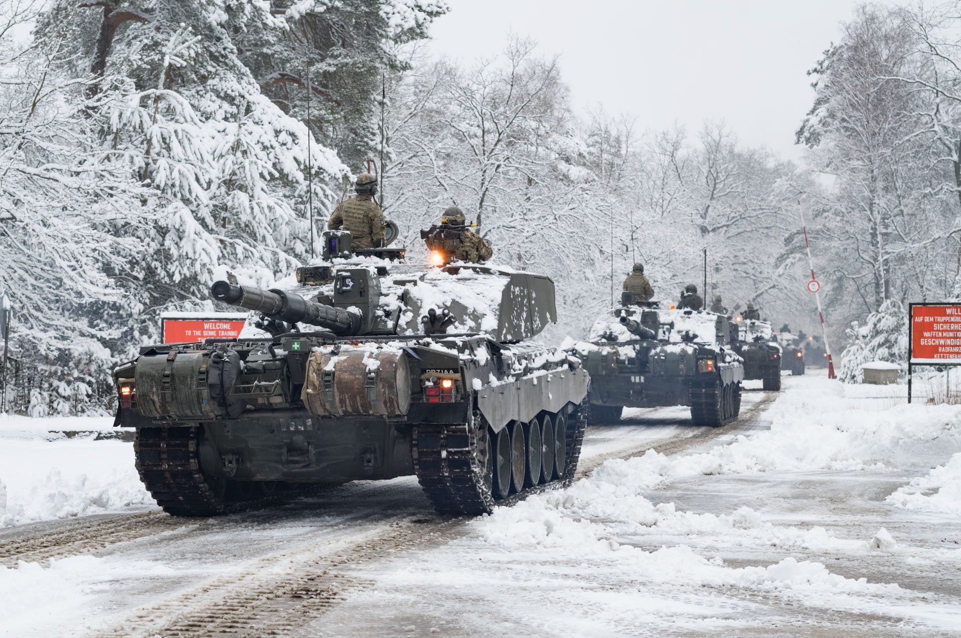 Tanks move through a snow-covered forest road with bare trees.
