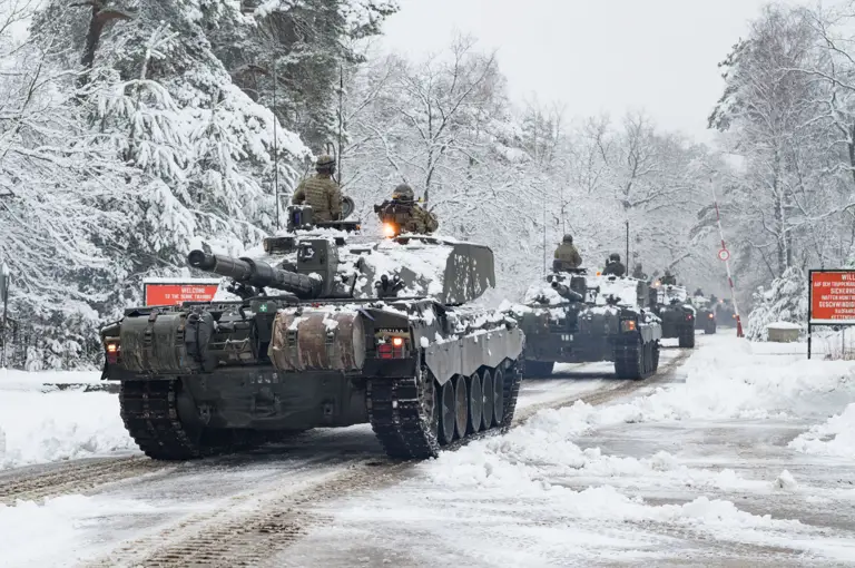 Tanks move through a snow-covered forest road with bare trees.