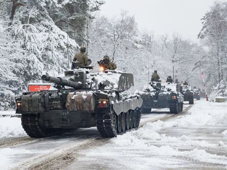 Tanks move through a snow-covered forest road with bare trees.