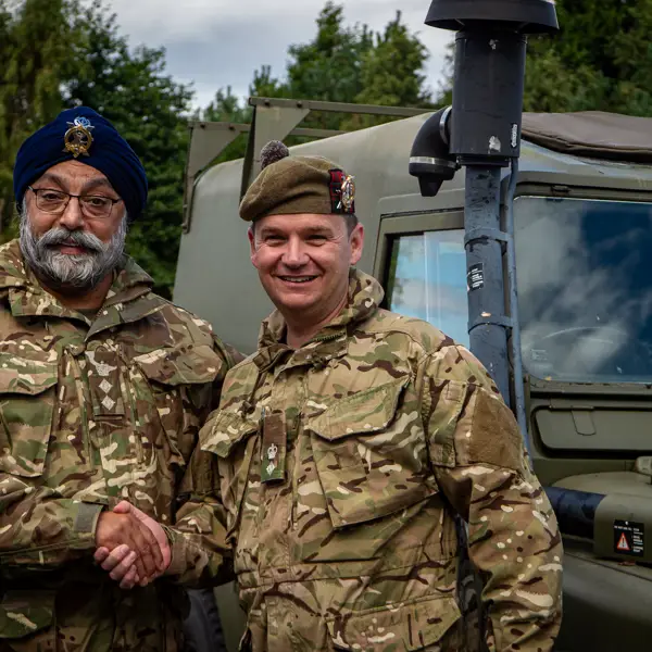 Two people in military uniforms are shaking hands in front of a military vehicle. One person is wearing a beret, and the other is wearing a turban with an emblem. Trees and a cloudy sky are visible in the background.
