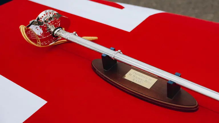 A sword is laid on a table, there is a Union Flag draped on the table underneath the sword.