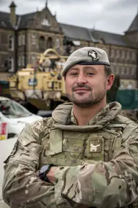 Soldier in camouflage uniform and beret stood posing for a portrait in front of a Jackal Vehicle