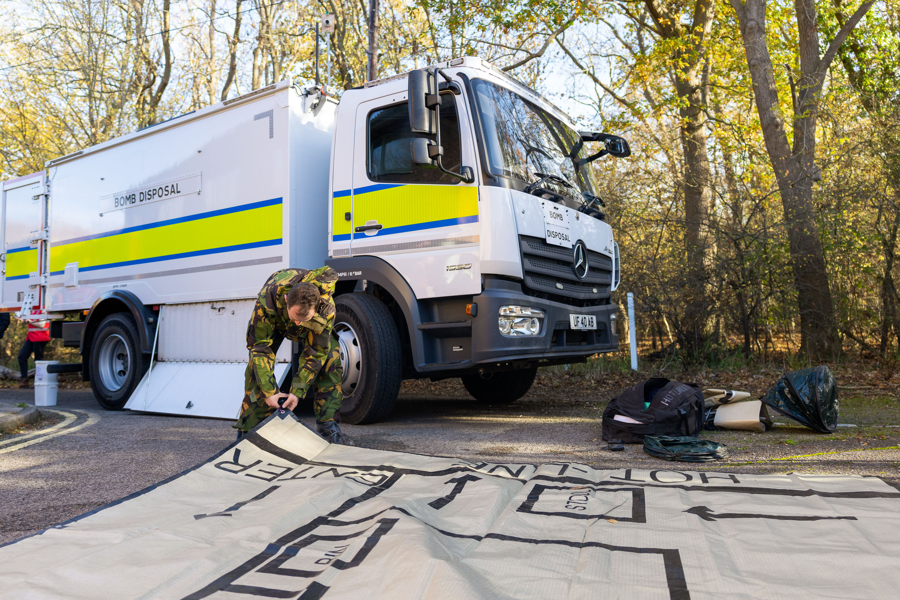 A soldier in a camouflage uniform rolls out a sheet of plastic in front of a white bomb disposal van.