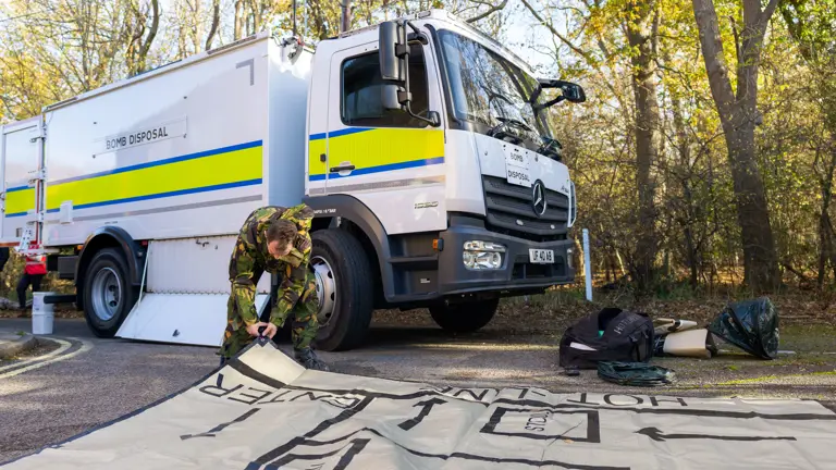 A soldier in a camouflage uniform rolls out a sheet of plastic in front of a white bomb disposal van.