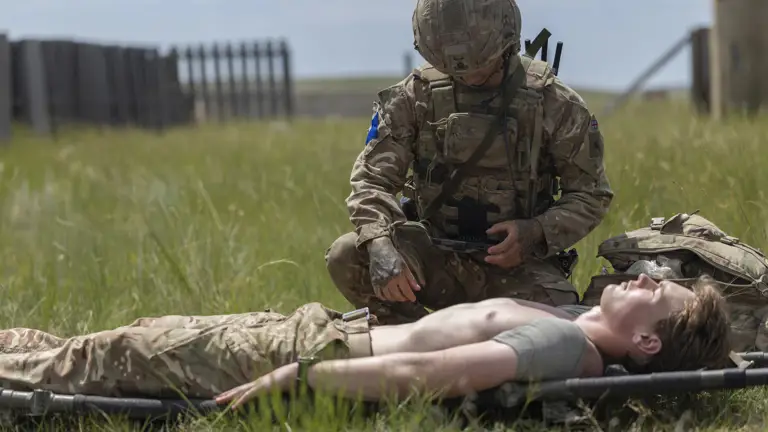 A person in military uniform tends to an individual lying on a stretcher in an open grassy field. There are structures visible in the background.