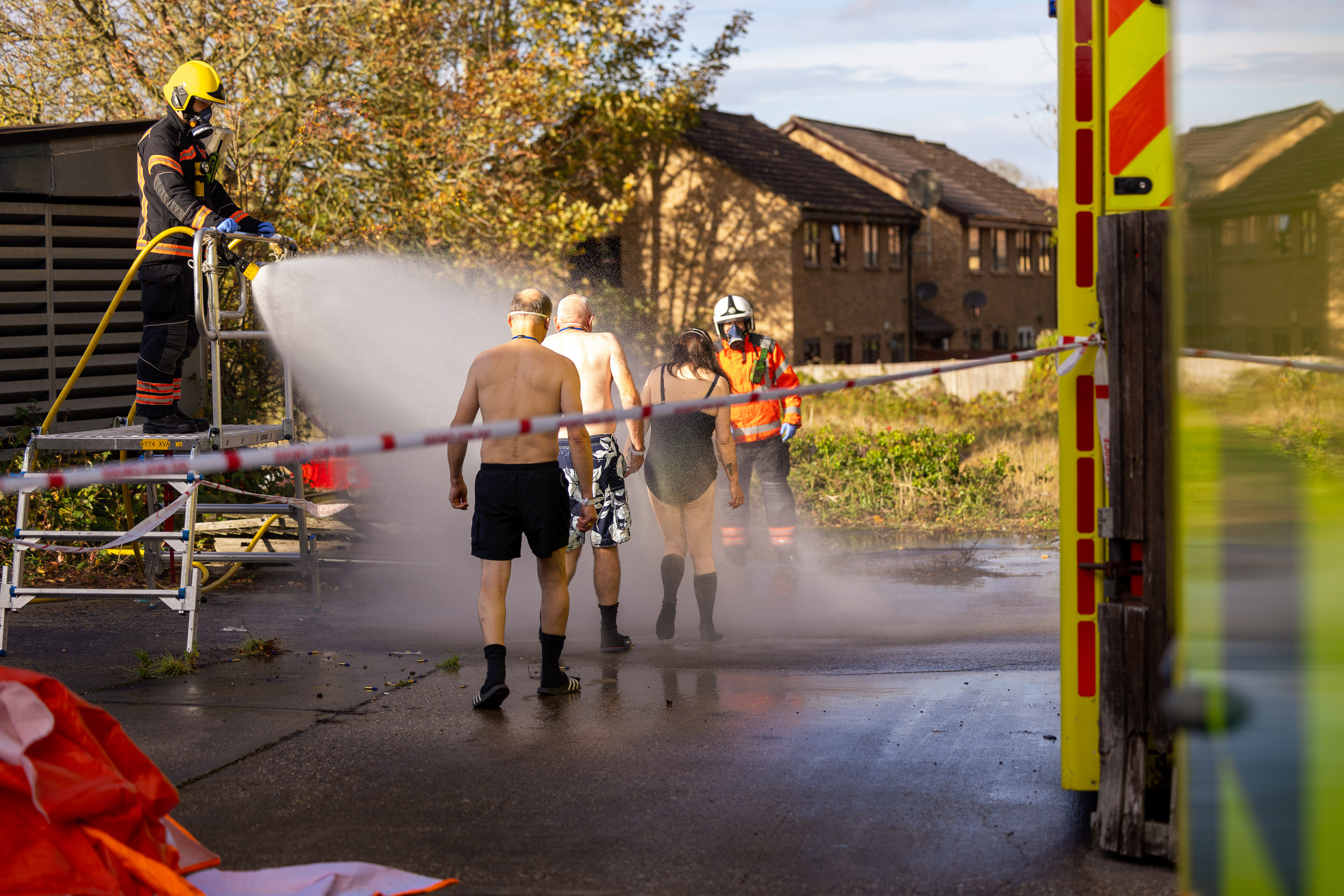 People in swimming costumes walk towards a firefighter. They are being sprayed by a hose.