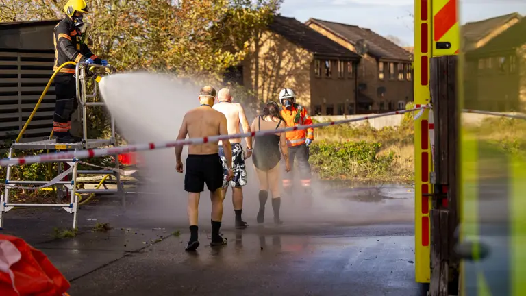 People in swimming costumes walk towards a firefighter. They are being sprayed by a hose.