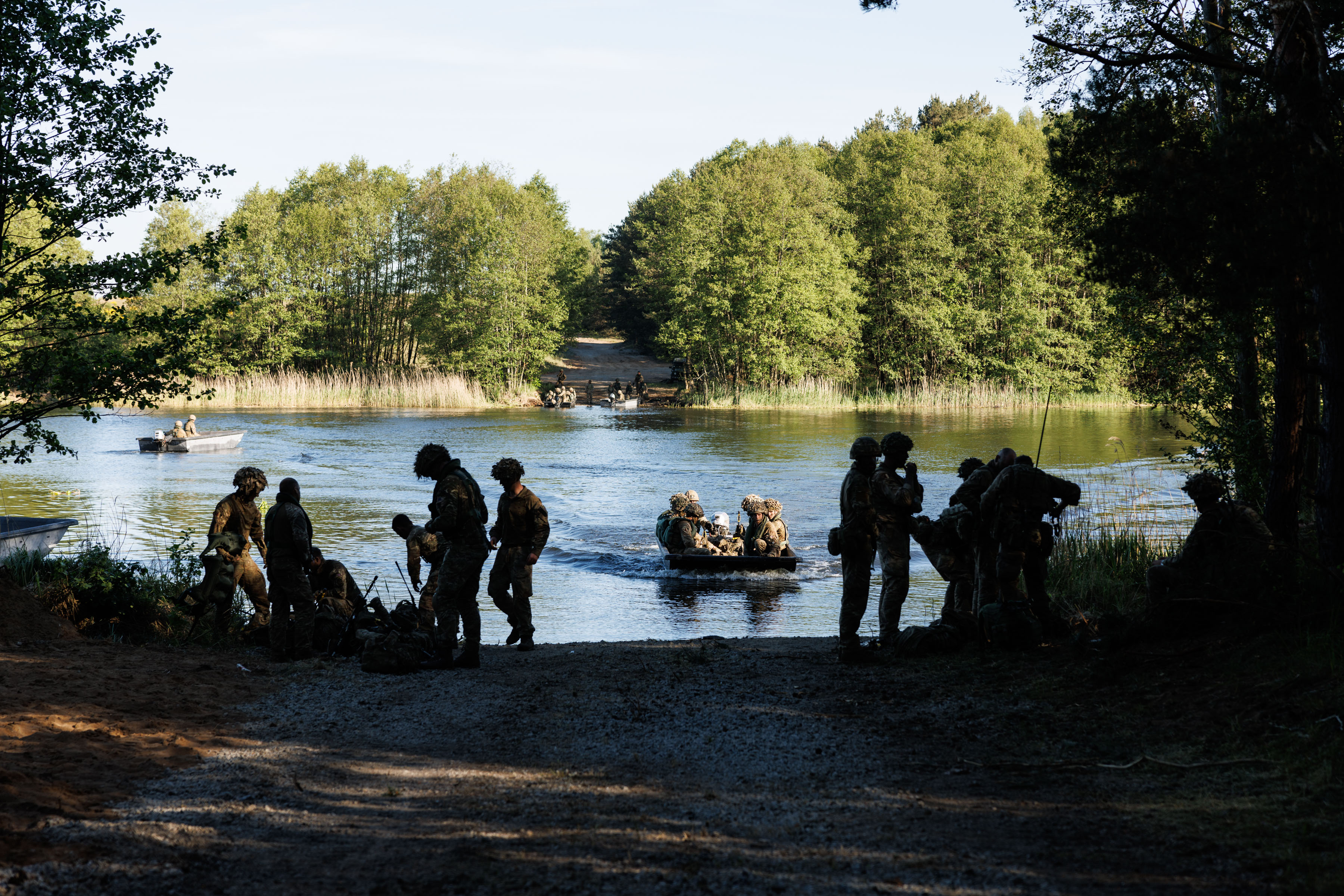 soldiers are seen crossing a river in rafts. the background shows a wall of trees and soldiers waiting to cross, soldiers and rafts are seen in the water and in the foreground, a opening in the wall of trees is where they are coming in to dock where soldiers can be seen sorting out their equipment. 