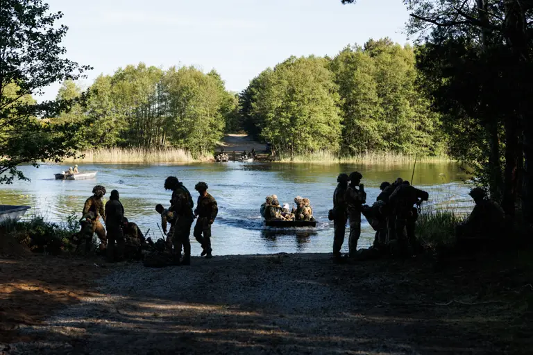soldiers are seen crossing a river in rafts. the background shows a wall of trees and soldiers waiting to cross, soldiers and rafts are seen in the water and in the foreground, a opening in the wall of trees is where they are coming in to dock where soldiers can be seen sorting out their equipment.