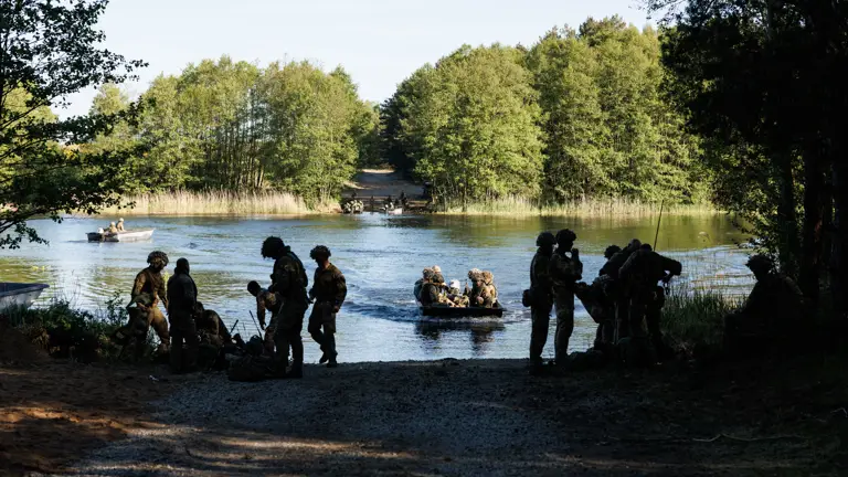 soldiers are seen crossing a river in rafts. the background shows a wall of trees and soldiers waiting to cross, soldiers and rafts are seen in the water and in the foreground, a opening in the wall of trees is where they are coming in to dock where soldiers can be seen sorting out their equipment.