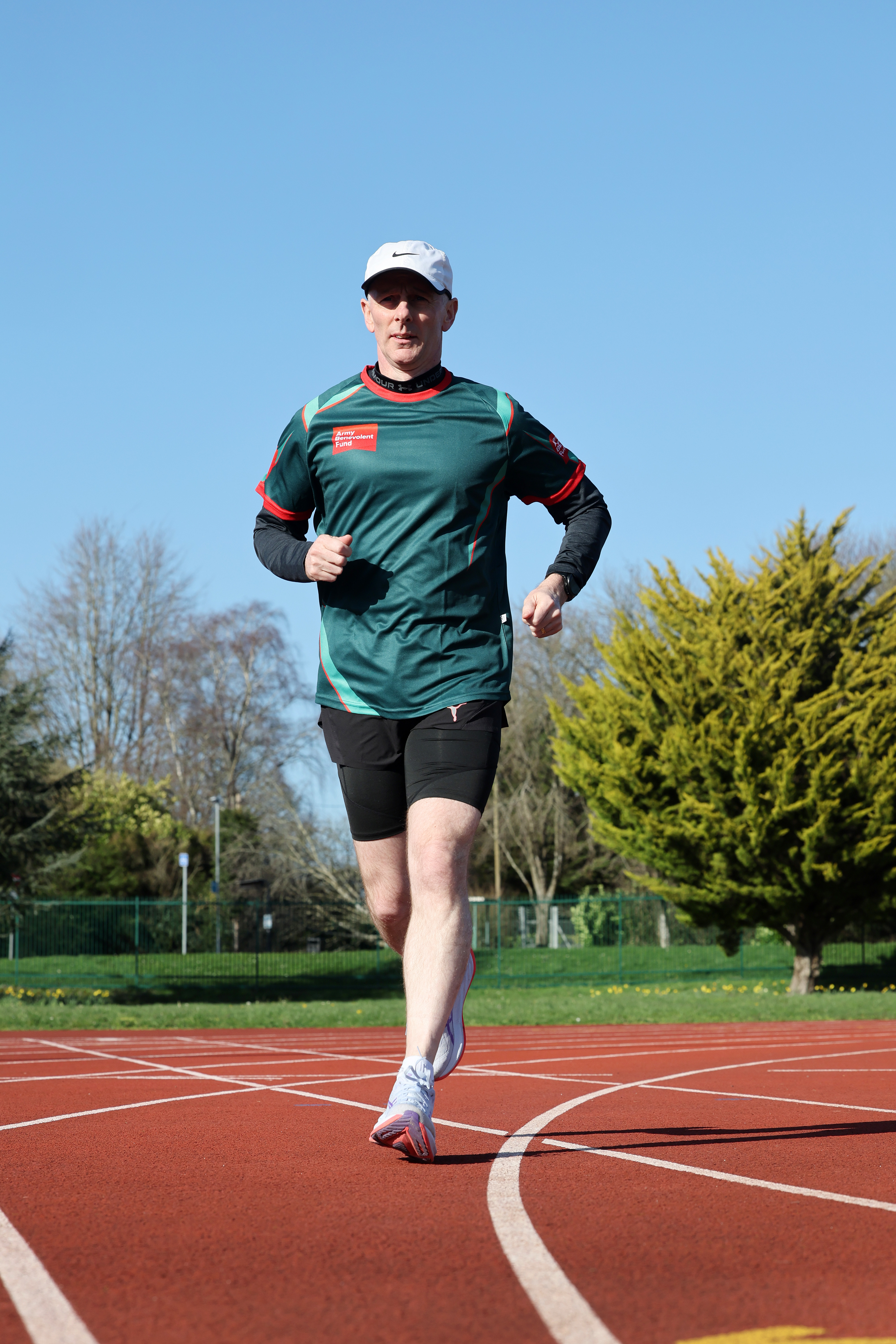 A man in athletic gear runs on an outdoor track under a clear blue sky. Green trees and a fence line the background, conveying a sense of focus and energy.