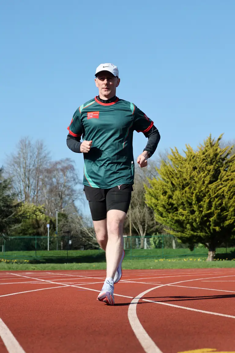 A man in athletic gear runs on an outdoor track under a clear blue sky. Green trees and a fence line the background, conveying a sense of focus and energy.