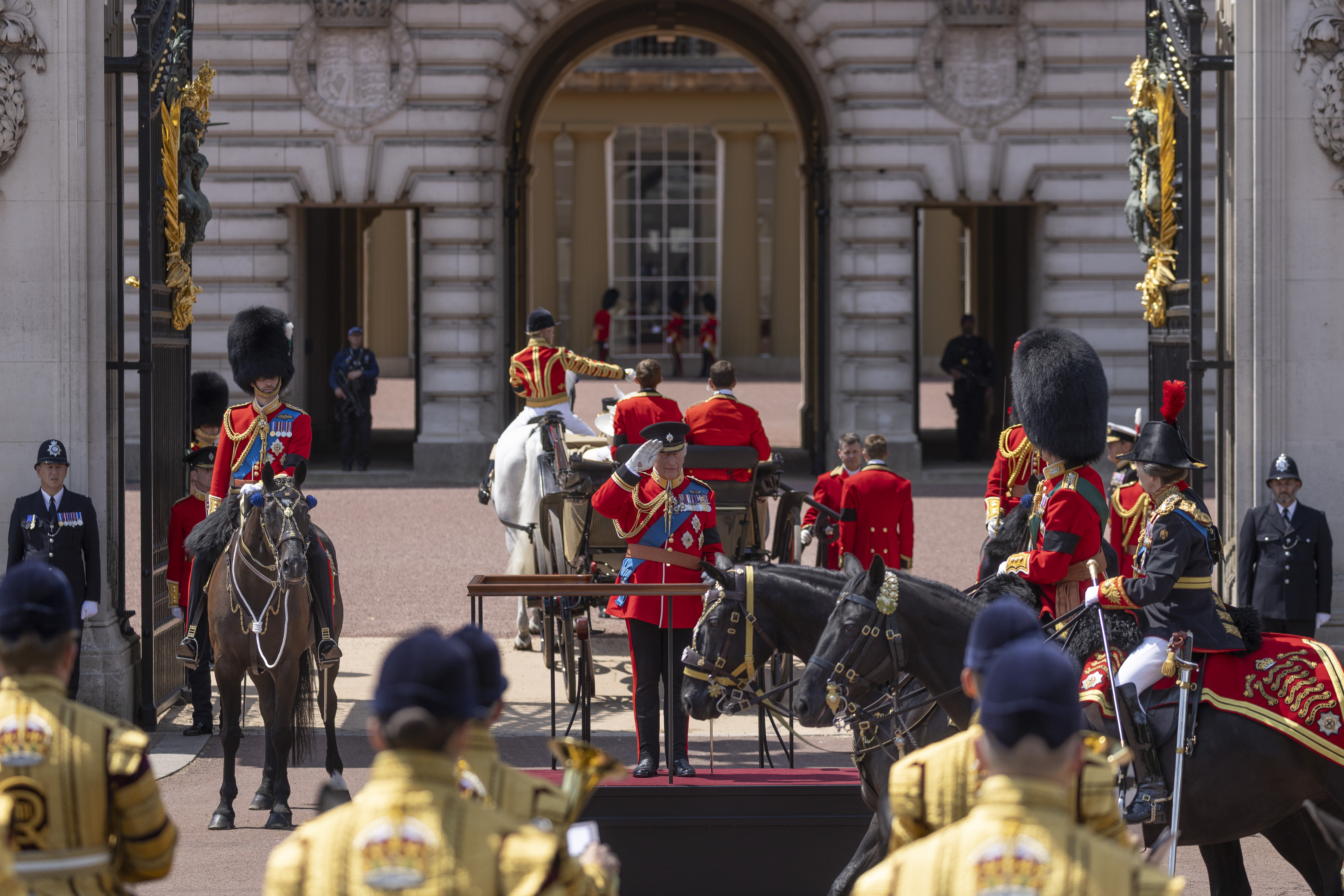 His Majesty The King salutes on a stage outside of Buckingham Palace as soldiers in ceremonial uniform look on.