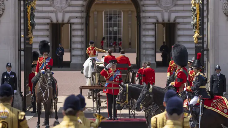 His Majesty The King salutes on a stage outside of Buckingham Palace as soldiers in ceremonial uniform look on.