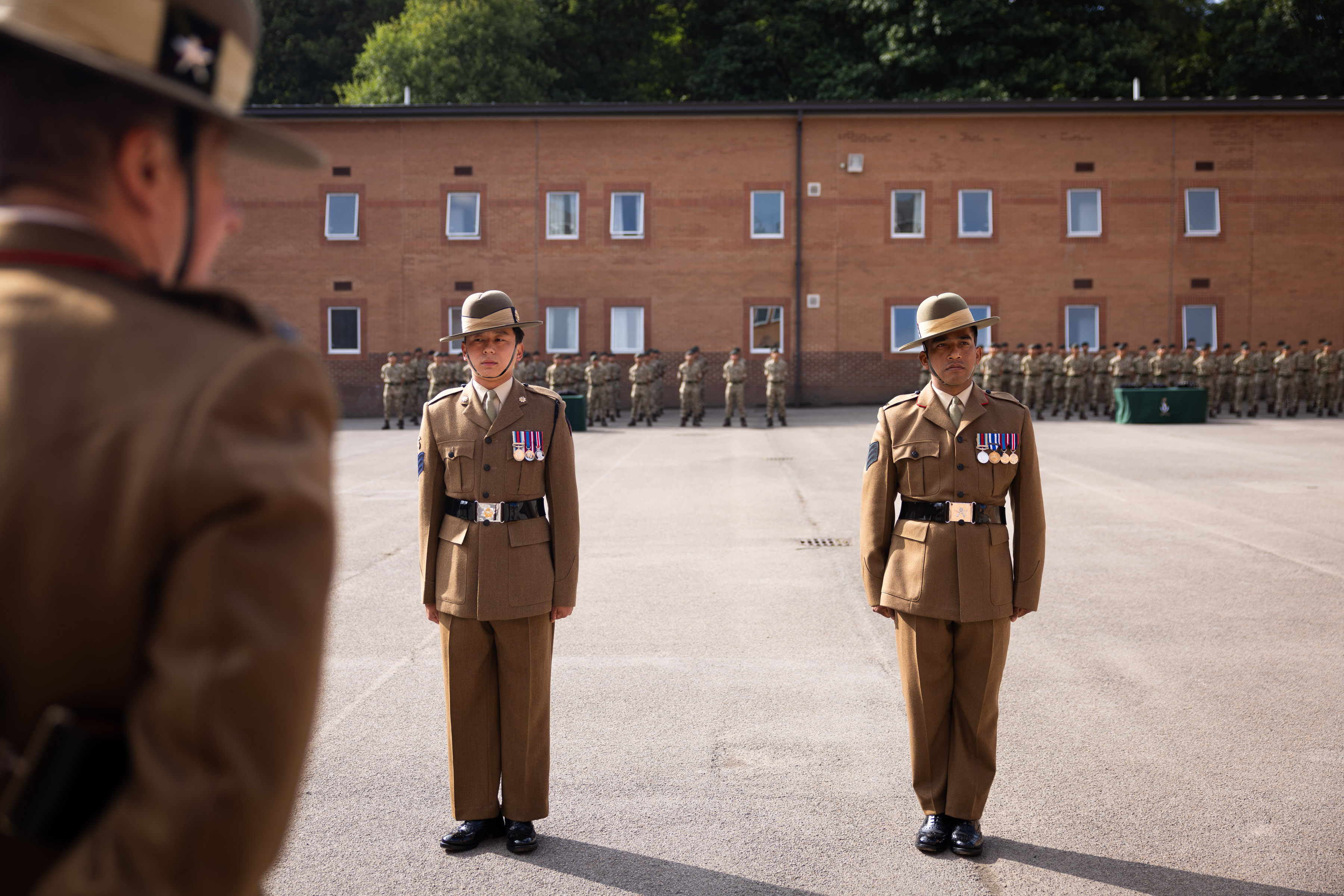 Two soldiers with brown smart uniforms and rows of medals across their chest stand in the middle of the parade square, there is another soldier in the foreground wearing brown uniform inspecting the two soldiers. The rows of soldiers in camouflage uniform are in the background. 