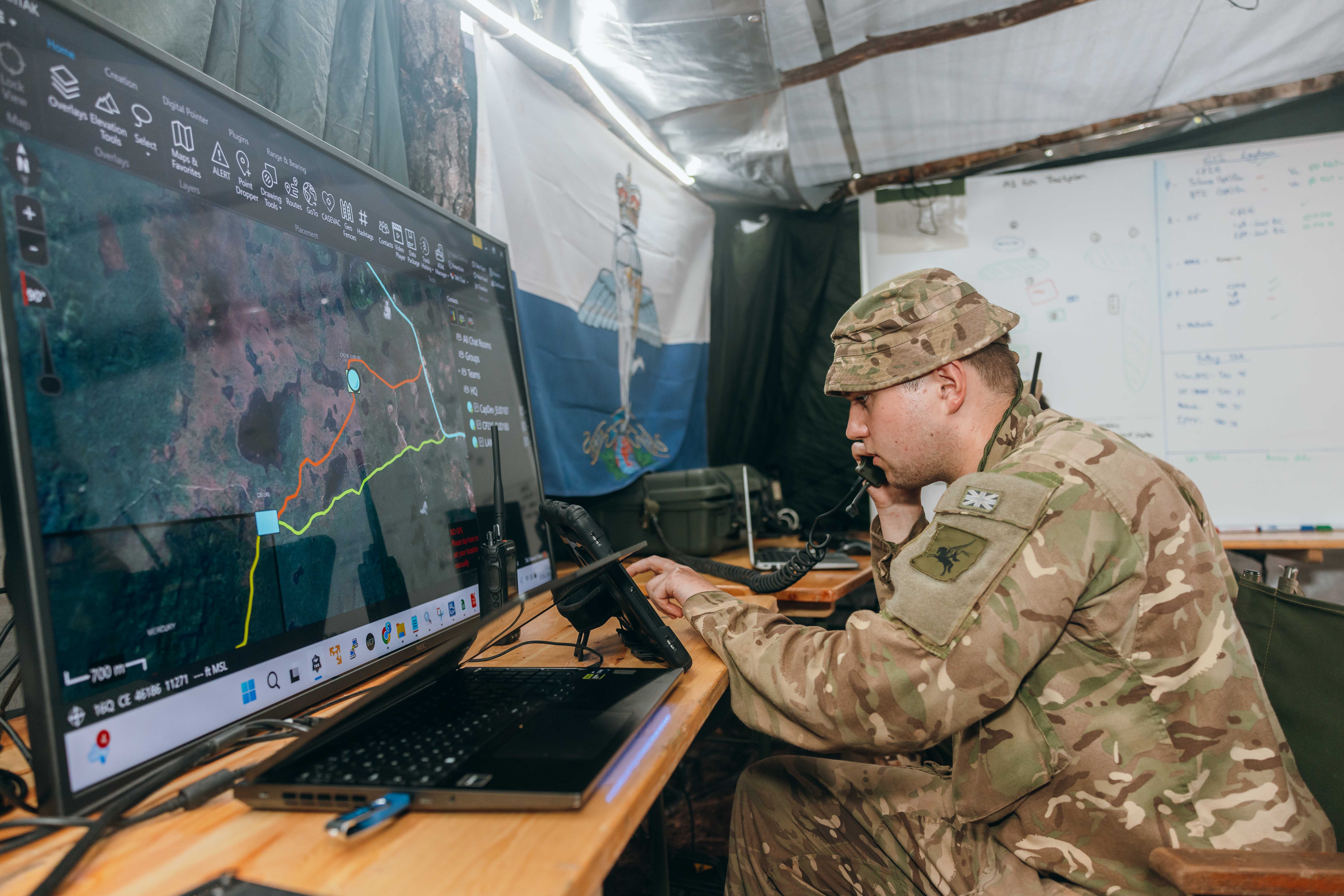 A soldier is sat at a desk in a military operations room. In front of him is a large display screen.