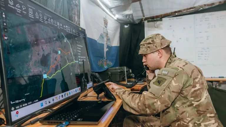 A soldier is sat at a desk in a military operations room. In front of him is a large display screen.