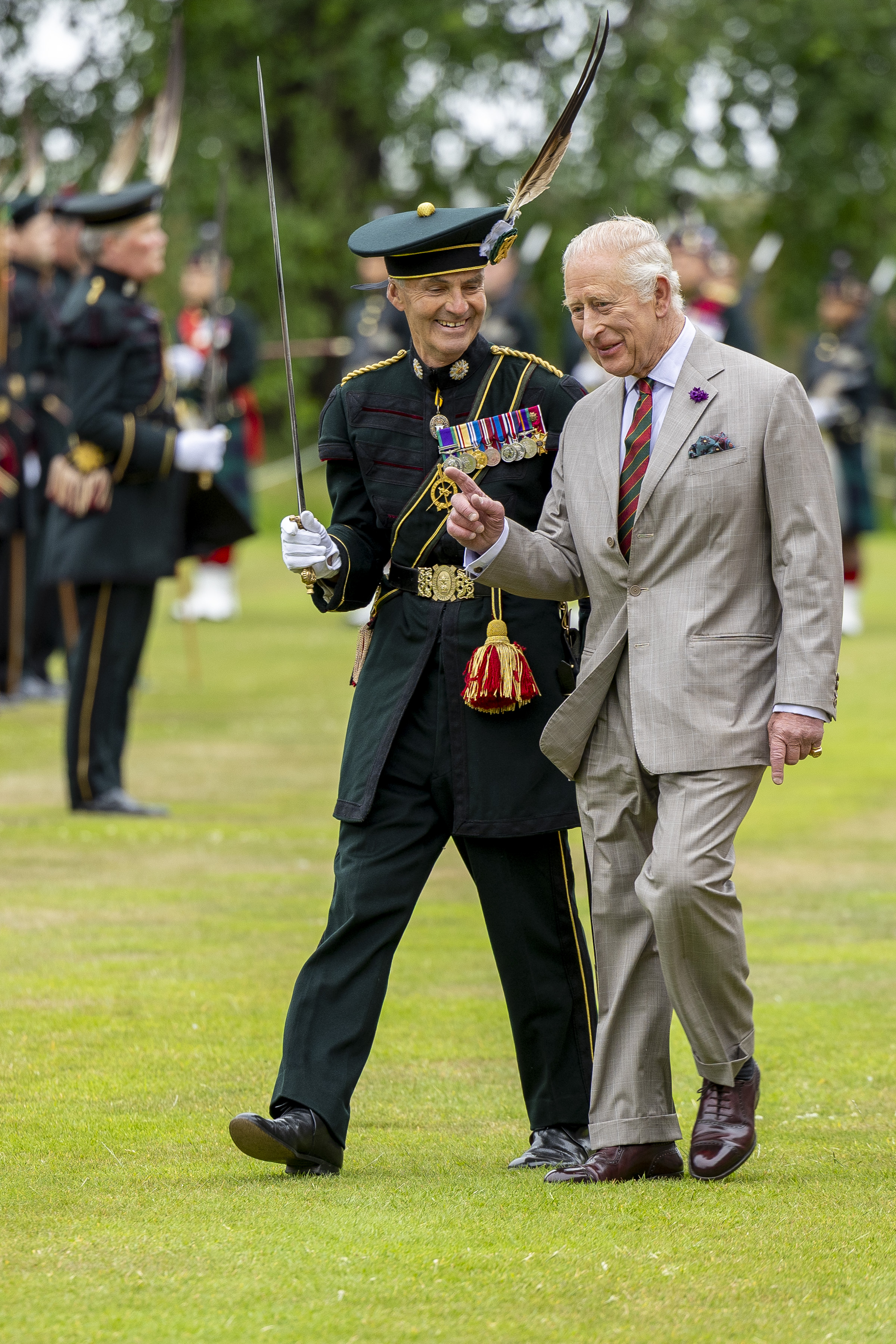 His Majesty The King is dressed in a suit with a thistle on the jacket, he is walking next to a soldier in ceremonial attire.
