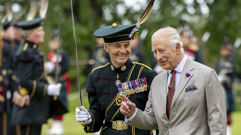 His Majesty The King is dressed in a suit with a thistle on the jacket, he is walking next to a soldier in ceremonial attire.