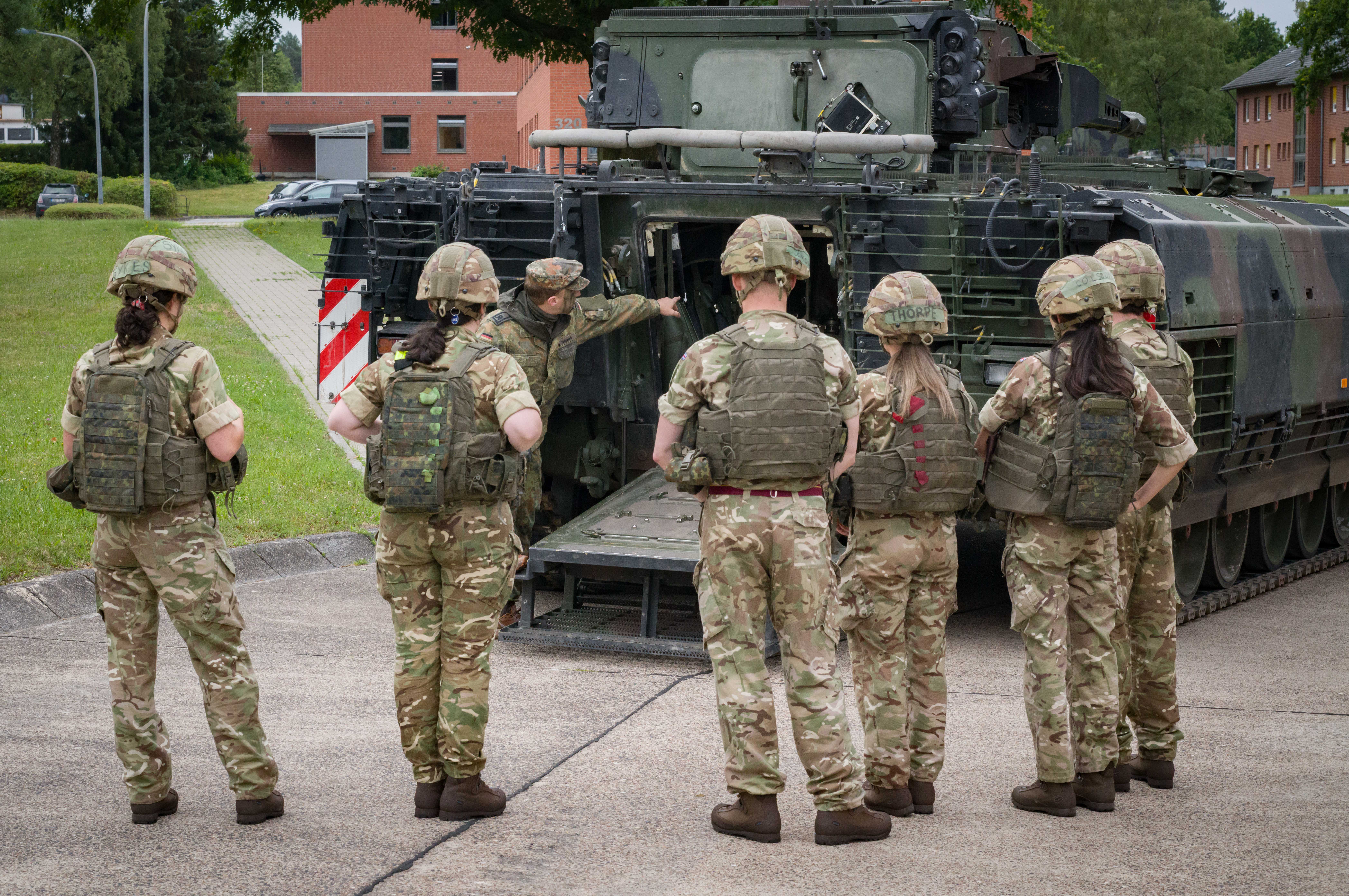 A group of military personnel in camouflage uniforms and helmets gather behind a military vehicle, possibly a tank, with one individual gesturing towards its open back. Buildings and trees are visible in the background.