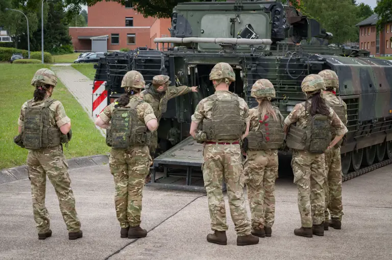 A group of military personnel in camouflage uniforms and helmets gather behind a military vehicle, possibly a tank, with one individual gesturing towards its open back. Buildings and trees are visible in the background.