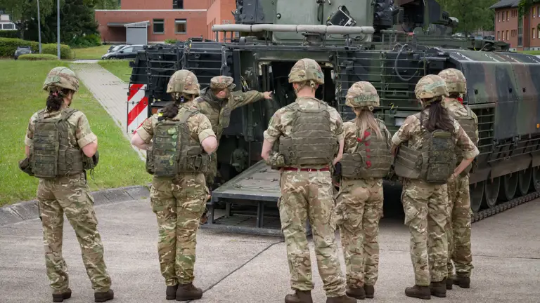 A group of military personnel in camouflage uniforms and helmets gather behind a military vehicle, possibly a tank, with one individual gesturing towards its open back. Buildings and trees are visible in the background.