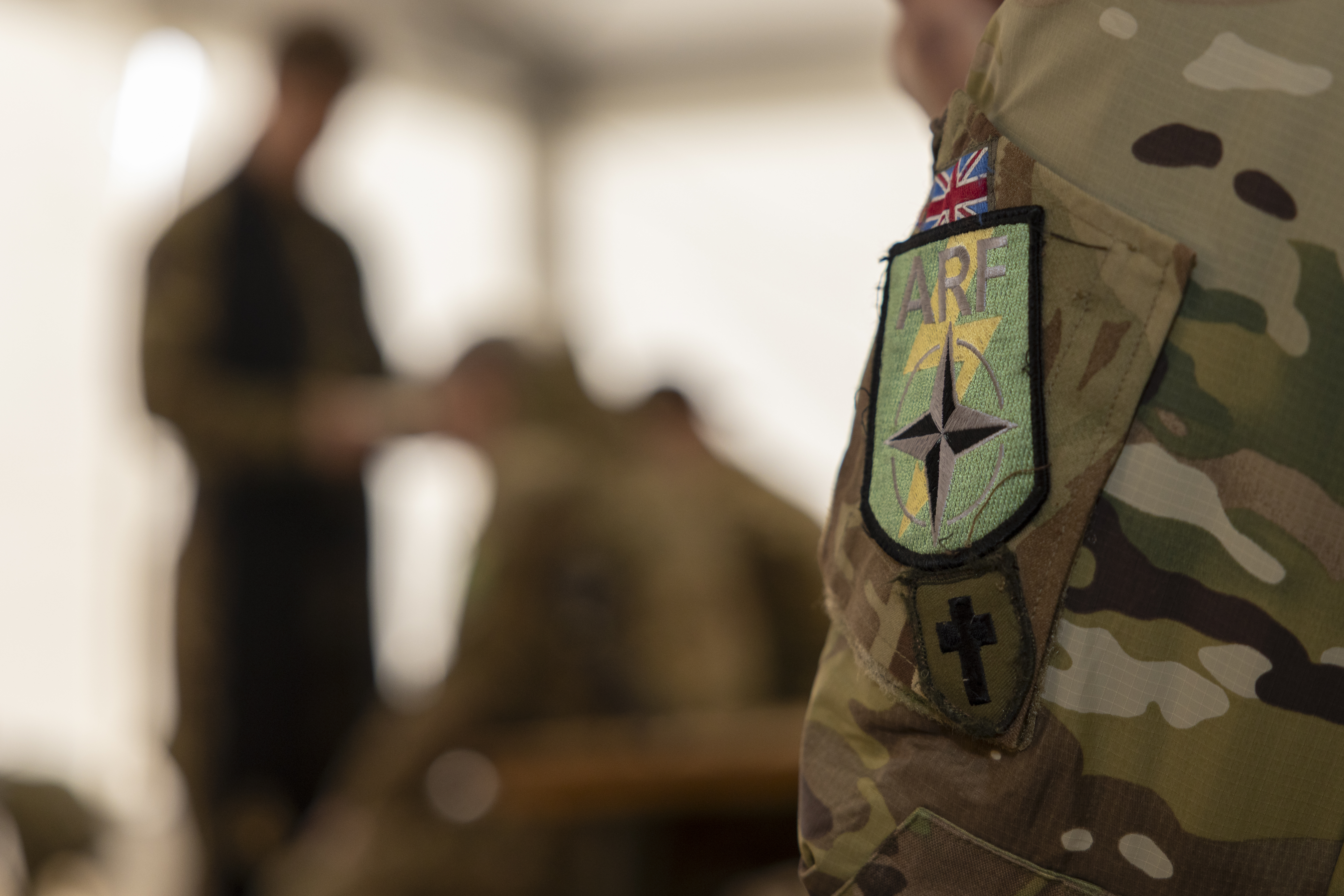 A close up of a British Army padre's shoulder, a British flag, a NATO ARF logo and a crucifix are located on the arm.