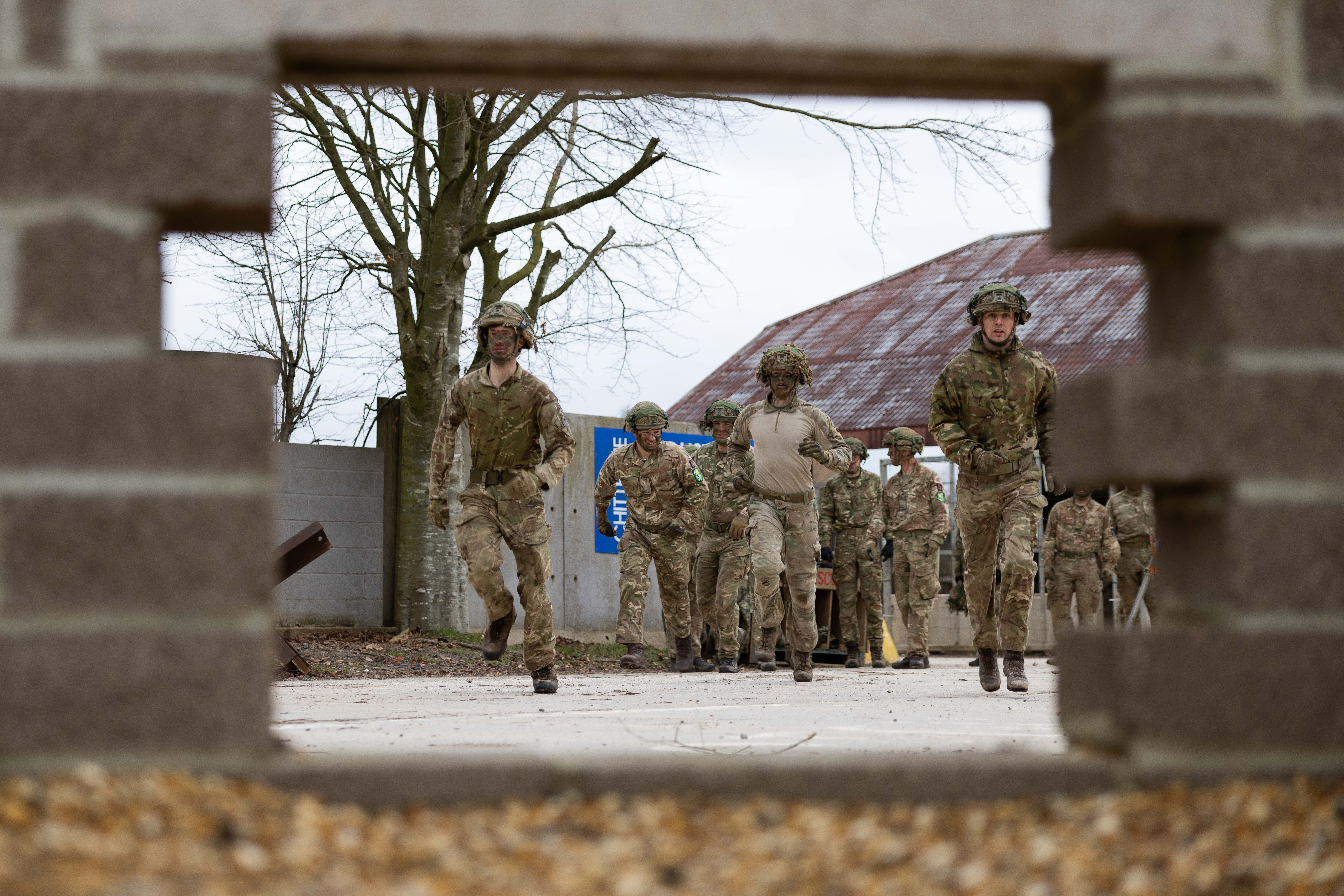 Multiple soldiers wearing camouflage uniform running at the start of an assault course training. Image is captured through a rectangle hole in a brick wall.