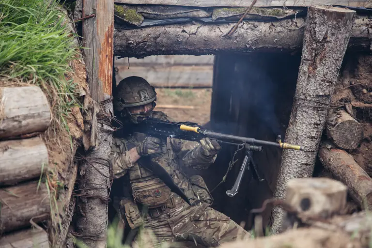 A soldier in camouflage uniform and helmet aims and fires his General Purpose Machine Gun during trench warfare training. 