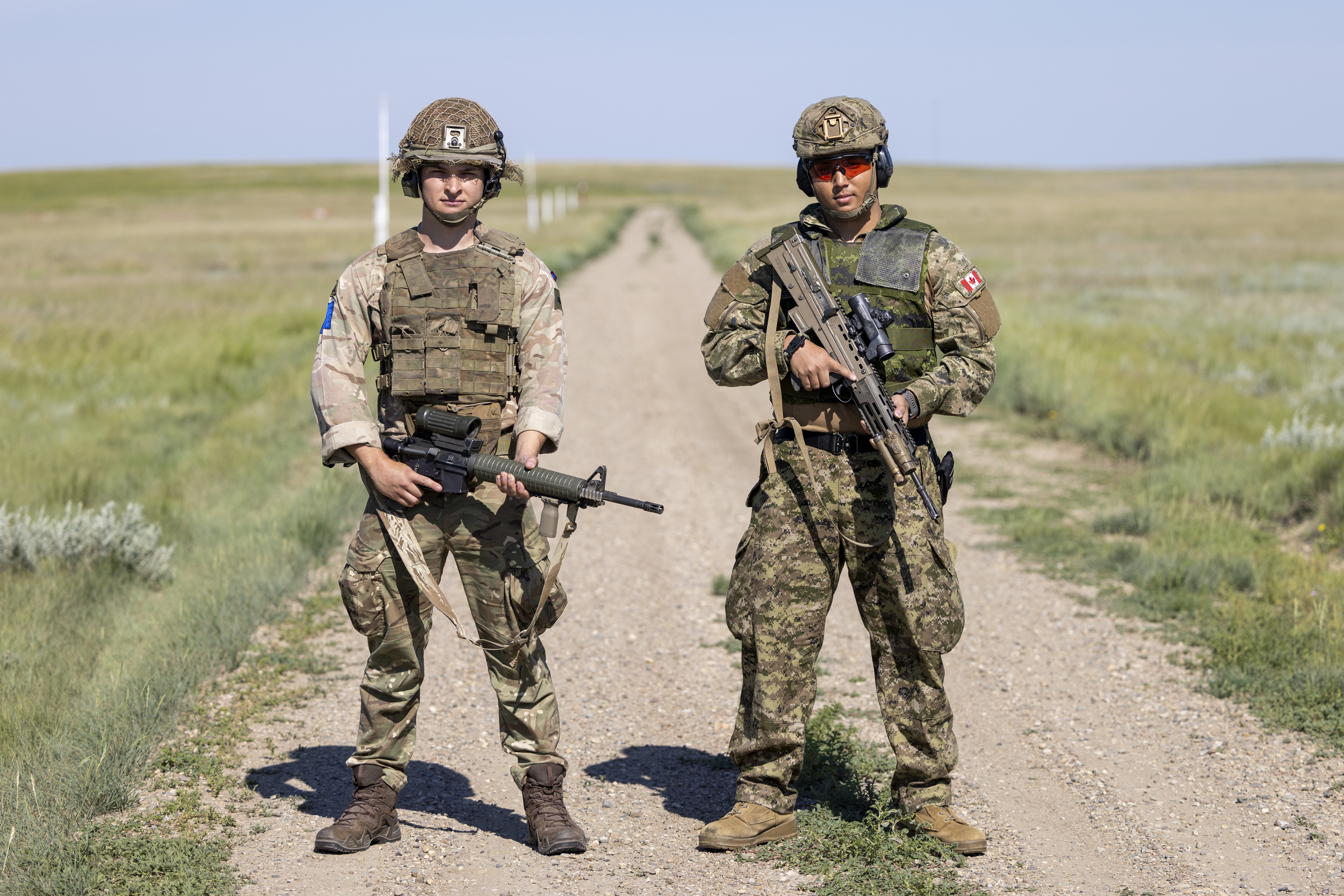 Two people in camouflage uniforms stand side by side. The uniform on the left features a Canadian flag and a Red Cross patch. The uniform on the right displays a UK flag patch and a badge with a stylized image.