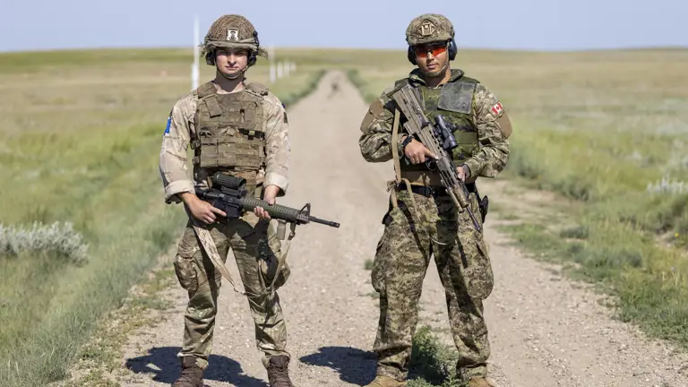 Two people in camouflage uniforms stand side by side. The uniform on the left features a Canadian flag and a Red Cross patch. The uniform on the right displays a UK flag patch and a badge with a stylized image.