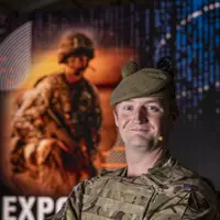 Soldier Stood in camouflage uniform and beret posing for a portrait in front of an Army Expo Poster