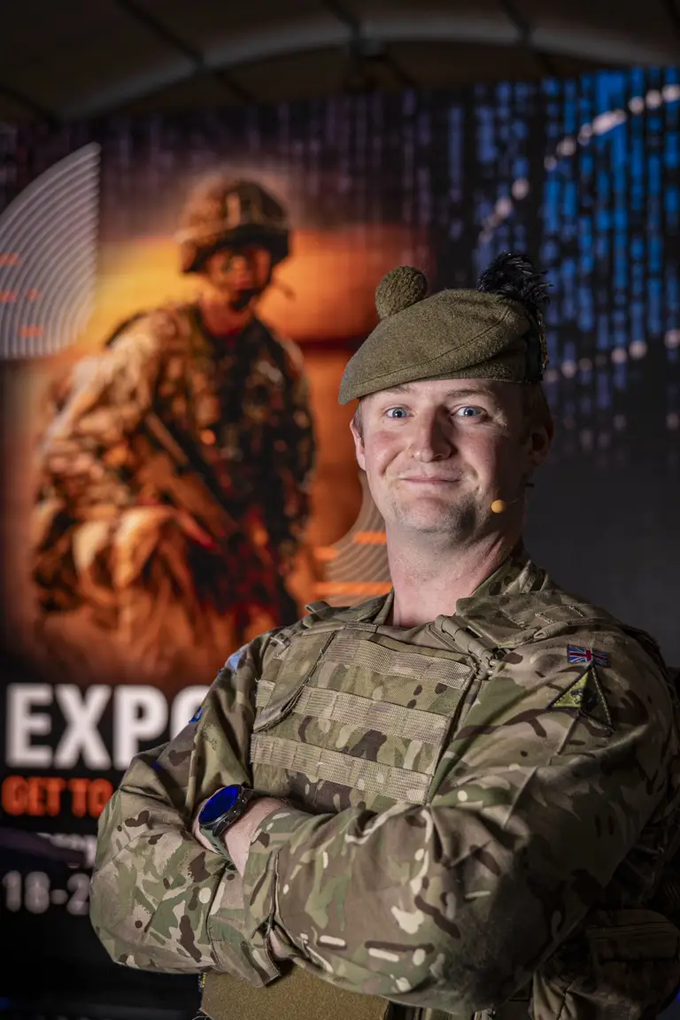 Soldier Stood in camouflage uniform and beret posing for a portrait in front of an Army Expo Poster