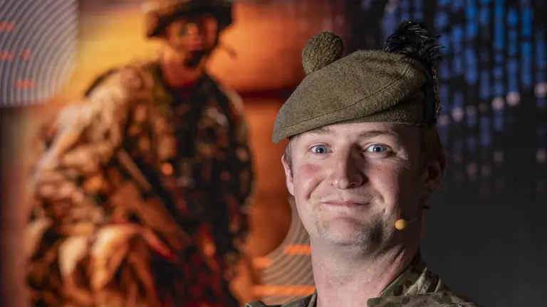 Soldier Stood in camouflage uniform and beret posing for a portrait in front of an Army Expo Poster