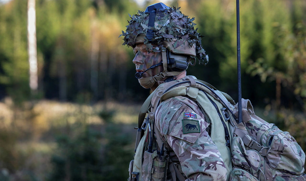 Soldier wearing camouflage uniform and helmet and body armour seen in the field, a bag on his back and an antenna coming out his bag and a microphone across his mouth.  