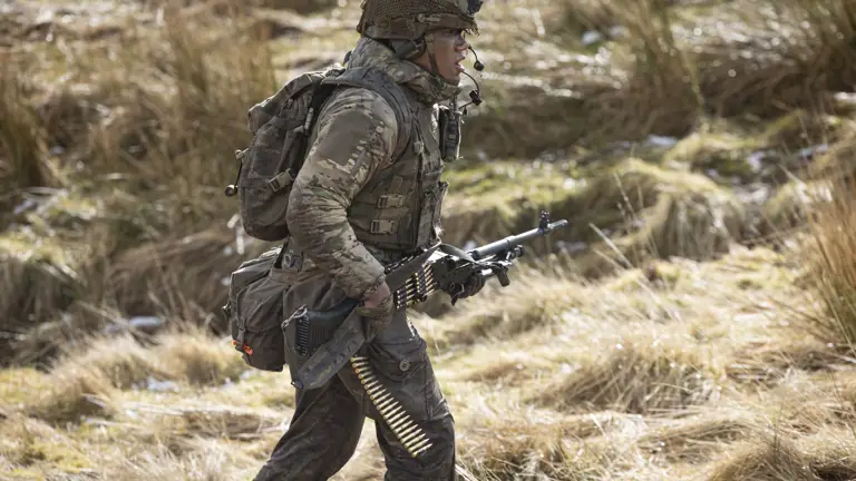 Soldier in camouflage uniform wearing body armour and helmet walks in the firing range holding his weapon